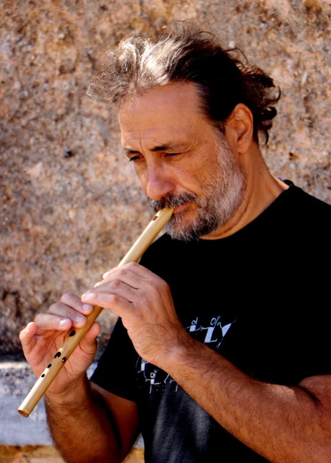 Musician playing a wooden flute in front of a stone wall.