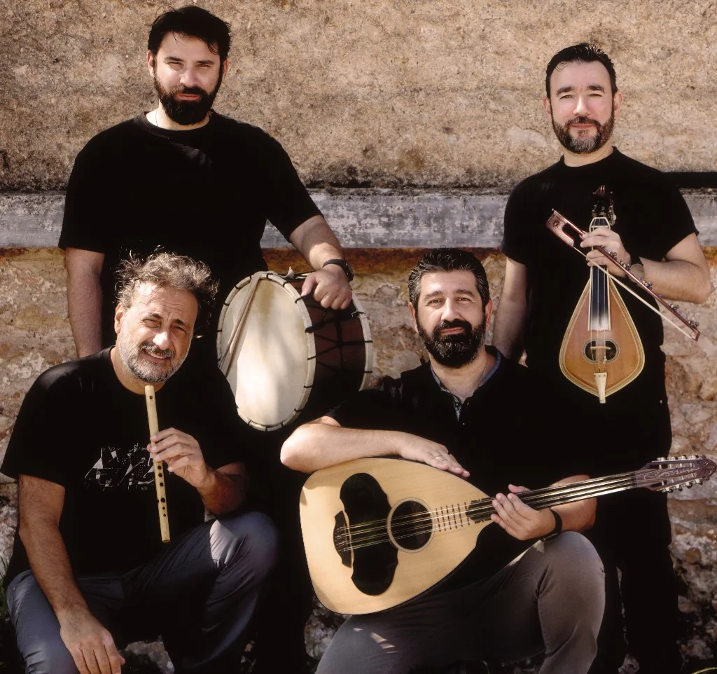 Esmiya: four musicians with lyra, frame drum, flute and laouto in front of a stone wall.