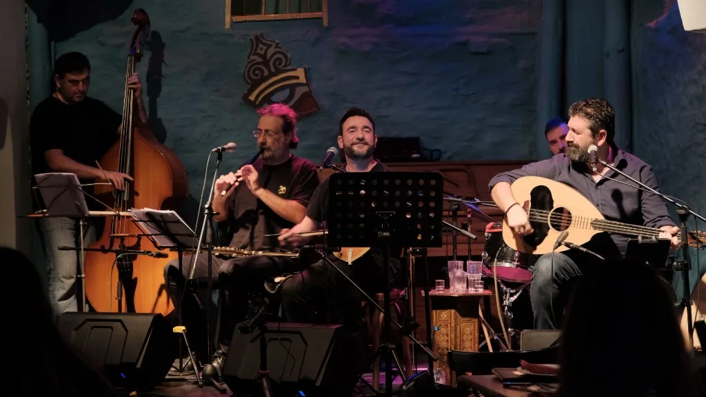 Four musicians performing live in front of a blue-lit stone wall.
