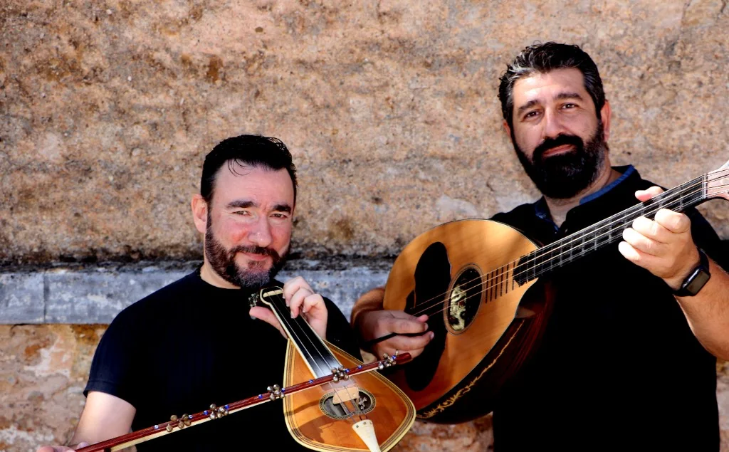 Two musicians with Cretan lyra and laouto in front of a stone wall.